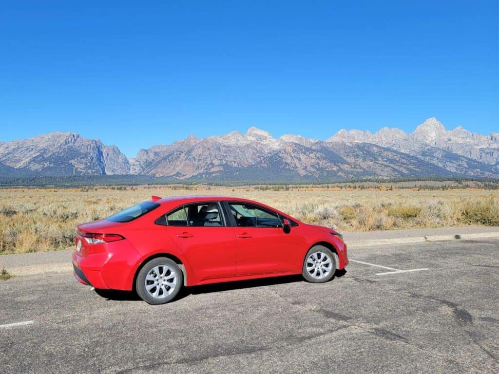 Red car parked on the side of the road with a row of mountains behind it.