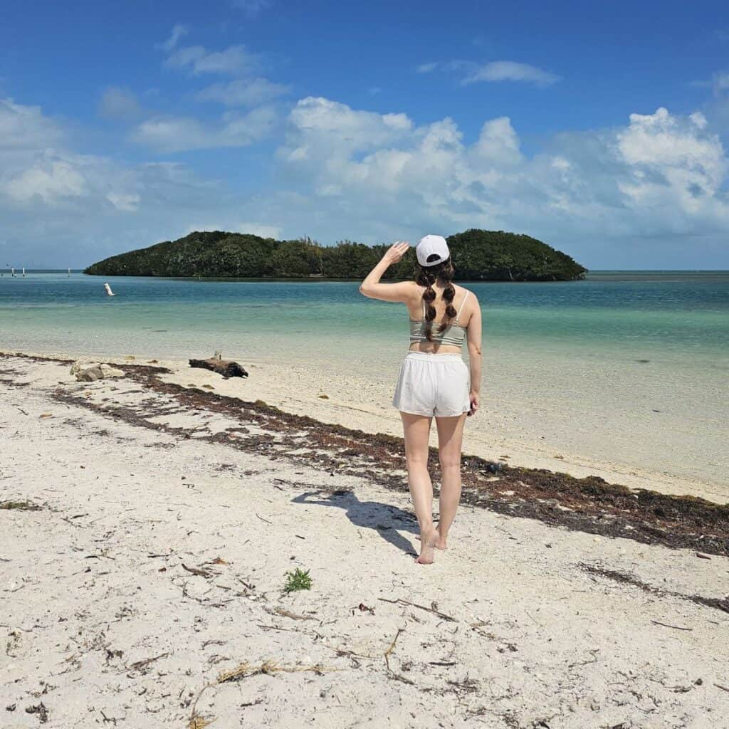 A woman with braids stands on a sandy beach looking out over the water in Biscayne National Park, Florida.